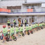 コース全体を引きで捉えた1枚。ヘルメットとビブスを着けた園児がペダルなし自転車を押して一列に並び、スタッフとくまるが見守っている